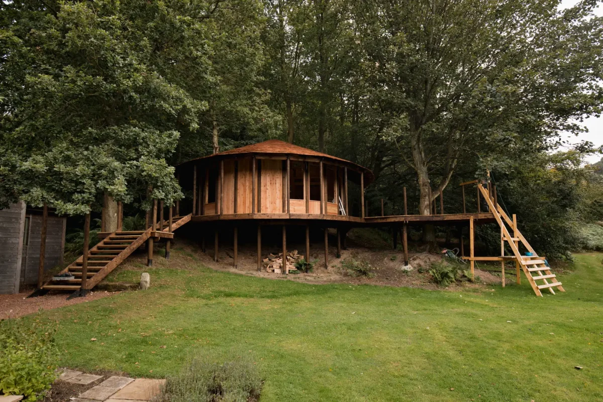 Cabane en bois perchée dans la forêt avec terrasse et escaliers, un hébergement insolite pour une expérience en pleine nature.