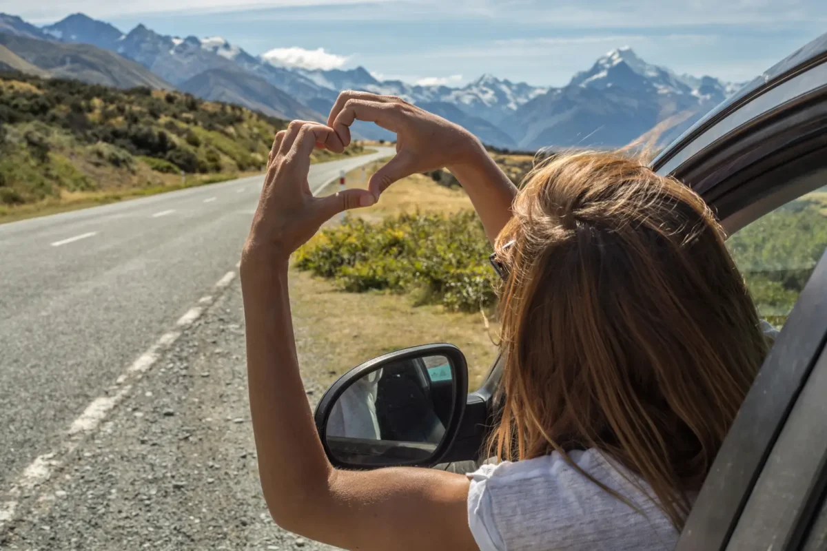 Voyageuse en road trip formant un cœur avec ses mains devant une route panoramique et des montagnes, symbole de liberté et d’aventure sur la route.
