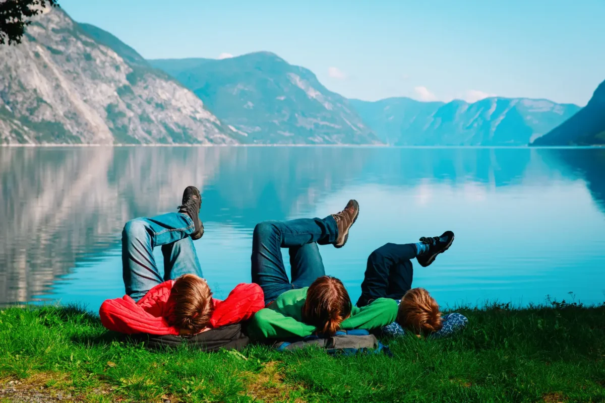 Trois voyageurs allongés sur l’herbe face à un lac paisible entouré de montagnes, profitant d’un moment de détente en pleine nature.